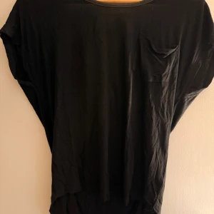 Svart topp med knappar från Mine - Svart loose-fit topp från Mine med korta ärmar och bröstficka. Baksidan har snygga knappar och dekorativa band i ryggen. Materialet är mjukt och lätt, perfekt för en chill och stilren look.