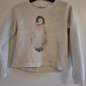 Vit fleece-tröja med pingvin från H&M - Supermjuk och fluffig vit fleece-tröja från H&M med lång ärm och en söt pingvin framtill. Perfekt för kalla dagar när du vill vara både varm och gullig. Tröjan har rund hals och ribbade muddar vid ärmslut och nederkant.
