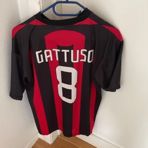 AC Milan Gattuso #8 matchtröja - Säljer en klassisk AC Milan fotbollströja med Gattuso och nummer 8 på ryggen. Tröjan har röda och svarta vertikala ränder, klubbmärke med stjärna på bröstet och är tillverkad i lätt polyester. Perfekt för dig som älskar italiensk fotboll och retrostil.