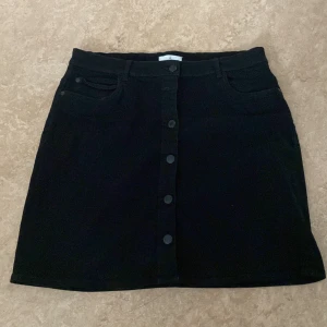 Svart jeanskjol med knappar - Svart kort jeanskjol med knappar framtill och klassiska fickor. Snygg och enkel modell som passar till allt, perfekt för dig som gillar en stilren look.