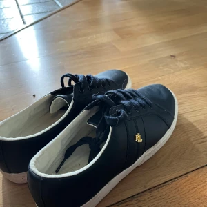 Mörkblå sneakers från Ralph Lauren - Stilrena mörkblå sneakers från Ralph Lauren med vit sula och guldfärgad RL-logga på sidan. Skorna har snörning och är gjorda i skinn med textilfoder. Använda 2 gånger och köpta för 750kr