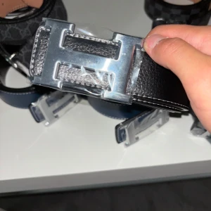 Svart bälte med H-spänne - Stilrent svart bälte från med ikoniskt spänne i silverfärgad metall. Bältet är gjort i präglat skinn och har en klassisk, rak form som passar till många olika outfits. Perfekt accessoar för att lyfta din look.
