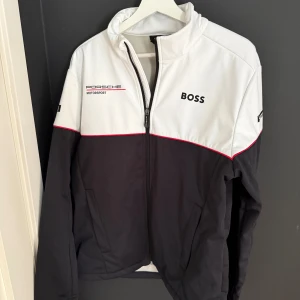 Porsche jacka - Snygg vindjacka från Porsche Motorsport och BOSS i svart och vitt med röda detaljer. Jackan har dragkedja, hög krage och loggor på bröst, rygg och ärmar. Perfekt för dig som gillar racing. Denna vara har slutat säljas nu och finns endast få kvar därav pris! ( ett par bränn märken på jackan men inget som syns jätte mycket )