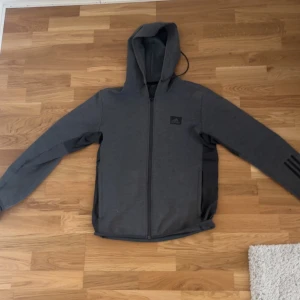 Grå hoodiejacka från Adidas M - Mörkgrå hoodiejacka från Adidas med dragkedja hela vägen, huva med snörning och svarta detaljer på ärmen. Mjuk bomullskänsla och klassisk Adidas-logga på bröstet. Perfekt för chill eller träning.