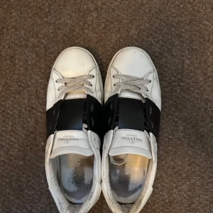 Valentino Garavani vita sneakers - Snygga vita sneakers från Valentino Garavani med bred svart läderdetalj över snörningen och ikoniska nitar på hälen. Skorna har rund tå, platt sula och klassisk siluett. Perfekta för dig som gillar exklusiva och trendiga sneakers.