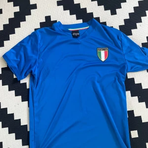 Blå Italien fotbollströja Infinity M - Snygg blå fotbollströja från Infinity i storlek M med Italiens emblem på bröstet och 'ITALIA' tryckt på ryggen. Tröjan har V-ringad krage med en randig detalj i italienska flaggans färger. Tillverkad i lätt och skönt syntetmaterial, perfekt för match eller träning.