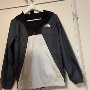 Svart och vit vindjacka från The North Face - Snygg vindjacka från The North Face i svart, grått och vitt. Jackan har huva, halv dragkedja framtill och elastiska muddar. Materialet är lätt och känns sportigt.