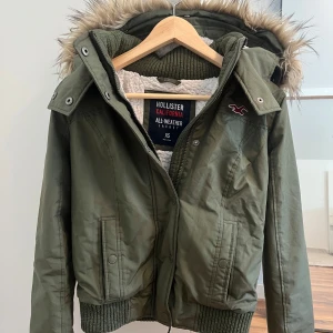 Grön Hollister bomberjacka - Snygg grön bomberjacka från Hollister med fuskpäls på luvan och mjukt vitt teddyfoder. Jackan har dragkedja, knappar och ribbade muddar. Perfekt för kalla dagar med sin varma insida och coola look. Klassisk Hollister-logga på bröstet. Säljer då jag inte för den till användning.