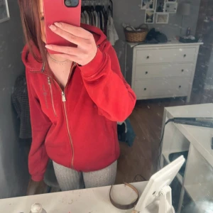 Röd fleecejacka från Sail Racing - Röd fleecejacka från Sail Racing med huva och dragkedja framtill. Jackan har en avslappnad passform och är perfekt för kyliga dagar. Mjuk och skön fleece på insidan, diskret logga på bröstet och praktiska fickor. Snygg och enkel att slänga på sig när det behövs lite extra värme.