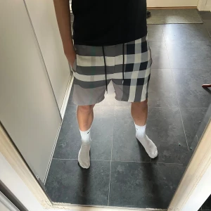 Burberry shorts  - Snygga Burberry shorts i klassiskt rutigt mönster med grå, svart och vit färg. Modellen har elastisk midja med svart snörning och metalländar med Burberry-logga. Materialet är mjukt och lätt, perfekt för sommaren. Fickor på sidorna för chill vibe.