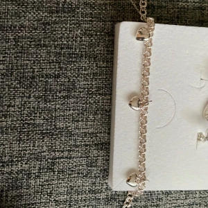 Silverfärgat armband med hjärtberlocker - Snyggt armband i silverfärg med små blanka hjärtberlocker som hänger längs kedjan. Kedjan har rundade länkar och ger ett stilrent och gulligt intryck. Perfekt för dig som gillar detaljer och vill ha något som sticker ut lite extra.