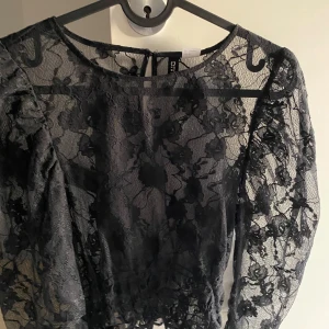 Svart spetsblus från Divided - Säljer en svart, transparent spetsblus från Divided med blommigt mönster. Blusen är croppad och har långa ärmar. Perfekt för dig som vill ha en edgy och trendig look med snygga detaljer.