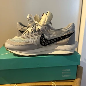 Nike x Sacai grå sneakers med mönstrad swoosh - Nike x Sacai sneakers i ljusgrått med mesh och mockadetaljer. Swoosh-loggan är svart med ett unikt mönster. Skorna har dubbla tungor, snörning och en chunky vit sula med rutmönstrad undersida. Perfekt för dig som gillar streetstyle och exklusiva samarbeten.