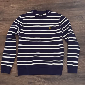 Randig tröja från Lyle & Scott, marinblå - Snygg marinblå tröja från Lyle & Scott med vita horisontella ränder och klassisk gul logga på bröstet. Tröjan har rund hals, långa ärmar och är gjord i mjuk bomull. Perfekt för dig som gillar stilren och sportig look.
