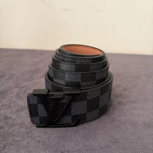 Louis Vuitton ruttig svart bälte - Snyggt Louis Vuitton-bälte med klassiskt damier-rutmönster i svart och grått. Bältet har en mattsvart LV-logga som spänne och är gjort i skinn med beige insida. Perfekt accessoar för att lyfta din outfit med en lyxig touch.