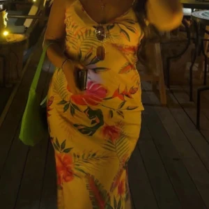 Lång blommig sommarklänning med slits - Snygg långklänning i ljusgul ton med tropiskt blommigt mönster i rött, grönt. Klänningen har smala axelband, v-ringning och en slits på sidan. Perfekt för sommarkvällar och semester vibes!