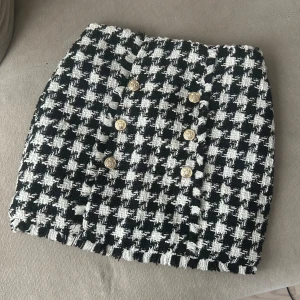 Svartvit tweed kjol med guldknappar - Elegant svartvit tweed kjol med guldknappar framtill. Kjolen har en diskret smal kedja på sidan. Aldrig använd, kommer med etiketter och extraknapp.