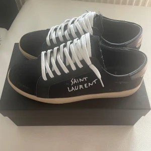 Svarta Saint Laurent sneakers med vit sula - Snygga svarta sneakers från Saint Laurent med vit sula och tydlig logga broderad i vitt på sidan. Skorna har vita snören och extra svarta skosnören medföljer. Ovandelen är i canvas och detaljer i skinn, perfekt för en clean och trendig look. Kan gå ner i pris vid snabbköp eller byten