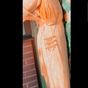 Beige kurdisk klänning med blommor - Superfin beige kurdisk klänning med vita blommor, v-ringad och med långa, vida ärmar. Klänningen har resår i midjan för en snygg passform och är gjord i ett lätt, flowy material. Perfekt för sommarens alla tillfällen! Storleken S och M✨