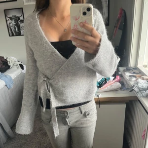 Grå omlottkofta från Gina Tricot - Supermjuk och mysig grå omlottkofta från Gina Tricot i ullblandning. Koftan har långa ärmar, v-ringning och knytband i sidan. Endast använd en gång.