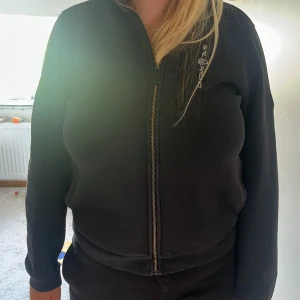 Svart hoodiejacka från Sail Racing - Svart hoodiejacka från Sail Racing med dragkedja framtill och broderad logga på bröstet. Jackan har huva, långa ärmar och är tillverkad i mjukt bomullsmaterial. Perfekt för chill dagar eller när du vill ha en sportig look.