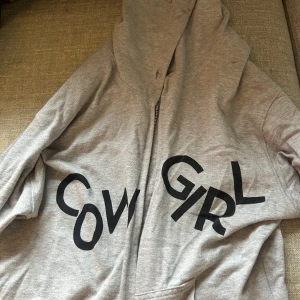 Vaquera Cowgirl grå hoodie med tryck - Grå hoodie från Vaquera med svart 'COWGIRL'-tryck över bröstet. Tröjan har huva med snörning, dragkedja framtill och känguruficka. Mjuk bomullskvalitet och långärmad modell med ribbade muddar. Perfekt statement-plagg för dig som gillar streetstyle. (AI)