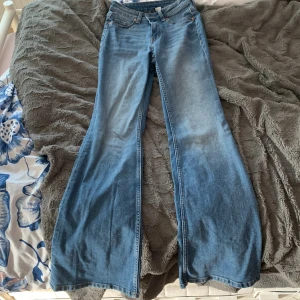 Blå bootcut jeans från H&M Divided - Säljer ett par ljusblå bootcut jeans från H&M Divided i storlek 36. Jeansen har klassisk femficksdesign, hög midja och vida ben med snygg utsvängning. Tillverkade i mjukt denimtyg med snygga slitningar framtill. Perfekta för dig som gillar retrovibbar! Och byxorna är tajta runt midjan.