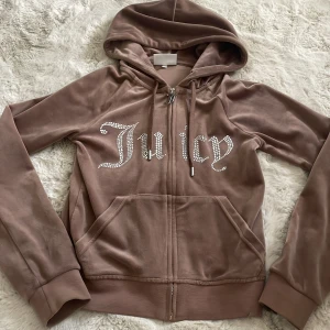Brun velour hoodie Juicy Couture XXS - Supermjuk brun velour hoodie från Juicy Couture i storlek XXS. Tröjan har huva, dragkedja och fickor framtill. På bröstet står det 'Juicy' i glittriga stenar. Perfekt för en chill och trendig look. Har tappat 1 glittrig Sten och har små bitmärken längst ner på tröjan (synns knappt).