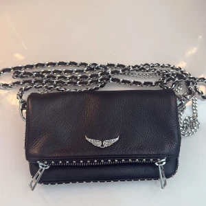 Svart axelväska Zadig & Voltaire - Cool svart axelväska i skinn från Zadig & Voltaire med silverfärgad kedjerem och ikonisk vingedetalj framtill. Väskan har dragkedja och små nitar längs kanterna för en edgy look. Perfekt storlek för det viktigaste och passar till många outfits. Använts en gång 