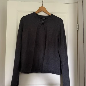 Mörkgrå kofta från Gina Tricot - Säljer en oanvänd stilren mörkgrå kofta från Gina Tricot. Koftan har rund halsringning, lång ärm och stängs med en knapp upptill. Den är stickad i mjukt material och har ribbade muddar vid hals och ärmslut. Storlek M!