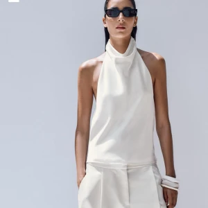 Vit halterneck linnetopp - Stilren vit halterneck topp med hög krage och ärmlös design. Toppens material har en lätt glans och faller snyggt mot kroppen, perfekt för en clean och modern look. Passar dig som gillar minimalistisk stil. Aldrig andvänd men ingen pris lapp kvar då jag tog bort den direkt efter köpet.💕💕