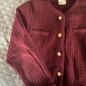 Vinröd stickad kofta med guldknappar - Vinröd stickad kofta med snygga guldknappar framtill och två dekorativa fickor på bröstet. Koftan har en grovstickad struktur och lång ärm, perfekt för lager-på-lager och en chill men ändå uppklädd vibe.Stär ej för postens slarv Köpt är köpt, gär ej att returneras. Pris går att diskuteras