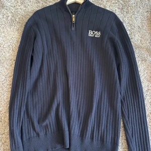 Mörkblå ribbad tröja från Hugo Boss - Snygg mörkblå ribbstickad tröja från Hugo Boss med halv dragkedja och broderad logga på bröstet. Tröjan har lång ärm, hög krage och är perfekt för dig som gillar stilren och klassisk design. Passar till jeans eller chinos för en clean look.