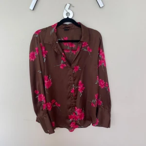 Brun blommig blus från Gina Tricot - Brun blus/ skjorta från Gina Tricot med rosa blommor och gröna blad. Skjortan har lång ärm, klassisk krage och knäppning framtill. Materialet känns mjukt och har ett lätt glansigt utseende. 