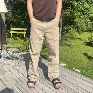 Beiga Carhartt Single Knee Pants 33/34 - Säljer ett par beiga Carhartt Single Knee Pants i 100% ekologisk bomull. Byxorna har loose passform, raka ben och flera praktiska fickor. Klassisk Carhartt-logga på bakfickan och robust känsla, perfekta för en chill och avslappnad stil.