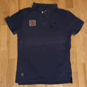 Svart piké från Morris - Snygg svart pikétröja från Morris i storlek S. Tröjan har klassisk krage, korta ärmar och två knappar framtill. Broderad logga på bröstet och en britiskflagg-patch ger en cool brittisk vibe. Tillverkad i 100% bomull för skön känsla.                                    KOLLA MINA ANDRA MORRIS ANNONSER!!