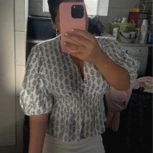 Vit och blå blus med puffärm - Supersöt blus i vitt med blått blommönster och puffiga korta ärmar. Smockad midja ger en snygg form och v-ringad halsringning gör den extra fin. Perfekt för vår och sommar när du vill ha en luftig och trendig look.