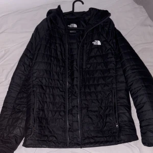 Svart dunjacka från The North Face - Svart dunjacka från The North Face med huva och dragkedja framtill. Jackan är quiltad och har två sidofickor med dragkedja samt en bröstficka. Perfekt för kyliga dagar och riktigt snygg streetstil.