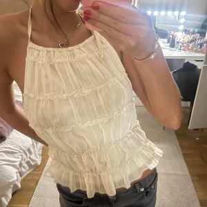 Vit volangig halterneck linnetopp - Supersöt vit halterneck topp med volanger och tunna knytband i ryggen. Toppen är ärmlös och har en luftig, rynkad design med flera volanglager 