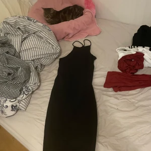 Svart långklänning med smala axelband - En stilren svart långklänning med smala axelband och figurnära passform. Klänningen är ärmlös och har en enkel, klassisk design som passar perfekt till många tillfällen. Materialet känns mjukt och följsamt mot kroppen.