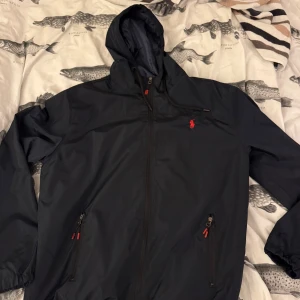 Svart vindjacka från U.S. Polo Assn XL - Snygg svart vindjacka från U.S. Polo Assn i storlek XL. Jackan har huva med dragsko, röda detaljer på dragkedjorna och klassisk röd logga på bröstet. Tillverkad i lätt polyester, perfekt för blåsiga dagar. Två fickor med dragkedja framtill. Den är även liten i storleken Så att den passar till folk som har M-L. Jag har storlek 5 och den är lite stor.