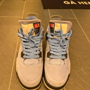 Nike Air Jordan 4 University Blue - Nike Air Jordan 4 University Blue med ljusblå mocka, meshpaneler och grå detaljer med svarta stänk. Klassisk siluett med snörning, svart och vit sula samt ikoniska Jumpman-loggan på plösen. Perfekta för dig som vill sticka ut med stilrena sneakers.