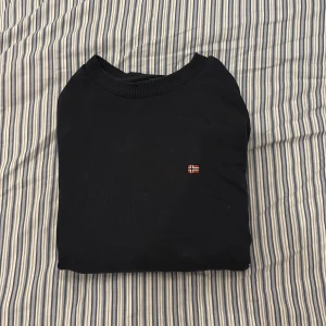 Svart sweatshirt från Napapijri - Svart/mörkblå sweatshirt från Napapijri med broderad norsk flagga på bröstet. Tröjan har rund hals, ribbade muddar och är långärmad. Perfekt basic med clean look och diskret branding, tillverkad i mjuk bomullsmix.