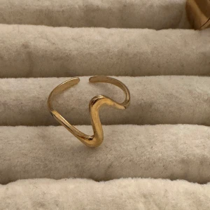 Guldig vågig ring i minimalistisk stil - Cool ring i guldton med vågig, öppen design. Perfekt för dig som gillar unika och trendiga smycken med minimalistisk känsla. Ringen är justerbar och har en snygg, modern form som sticker ut.