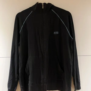 Svart zipjacka från Hugo Boss - Svart zipjacka från Hugo Boss med diskret logga på bröstet och ljusblå detaljer längs axlarna. Jackan har huva, hel dragkedja och två fickor framtill. Perfekt för en chill och stilren look.