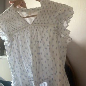 Vit blus med blått mönster Gina Tricot - Supersöt vit blus från Gina Tricot med blått diskret mönster. Blusen har volangdetaljer vid ärmarna och v-ringad hals. Tunn och luftig i bomull, perfekt för varma dagar. Säljs i storlek 158/164.
