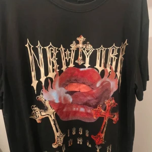 Svart t-shirt med tryck från Romwe - Cool svart t-shirt från Romwe med stort tryck framtill: röda läppar, kors och texten 'New York'. Oversized vibe och korta ärmar, perfekt för dig som gillar statement-plagg. Materialet känns mjukt och skönt mot huden.