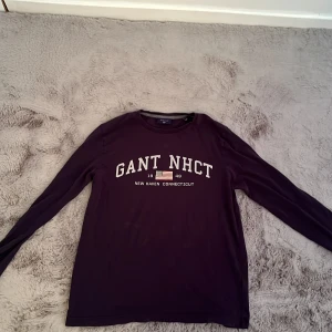 Mörkblå långärmad tröja från Gant - Säljer en mörkblå långärmad tröja från Gant med tryckt text 'GANT NHCT' och amerikansk flagga framtill. Tröjan har rund halsringning och är i mjuk bomull, perfekt för en chill och stilren look.