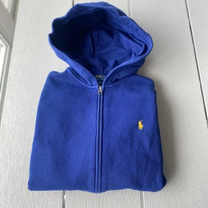 Blå hoodie – Polo Ralph Lauren – Strl XS - Färgstark blå hoodie från Polo Ralph Lauren, storlek XS (160/80). 💛 Gult broderat Polo-emblem 🧥 Dragkedja framtill 💙 Sitter snyggt – tajtare passform 📍 Mycket bra skick  Perfekt för dig som gillar klassisk stil med färg.