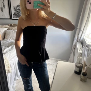 Svart axelbandslös tubtopp med volang - Säljer en svart axelbandslös tubtopp med snygg volang nertill. Toppen är figurnära och har en peplum-liknande form som ger en cool siluett. Perfekt att styla med jeans eller kjol för en trendig look.